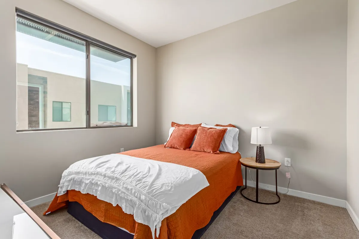 A cozy bedroom featuring a bed with an orange bedspread and pillows, a nightstand with a lamp, and a large window allowing soft, natural light to fill the room. The mood is serene and inviting, perfect for restful nights and peaceful mornings.
