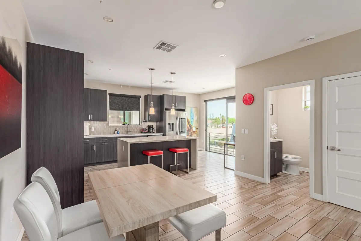 A bright, open-plan interior featuring a kitchen island with red bar stools, a dining table, and expansive large windows that flood the space with sunlight. The generous glazing on two sides offers sweeping views and creates a seamless connection between living and dining areas.