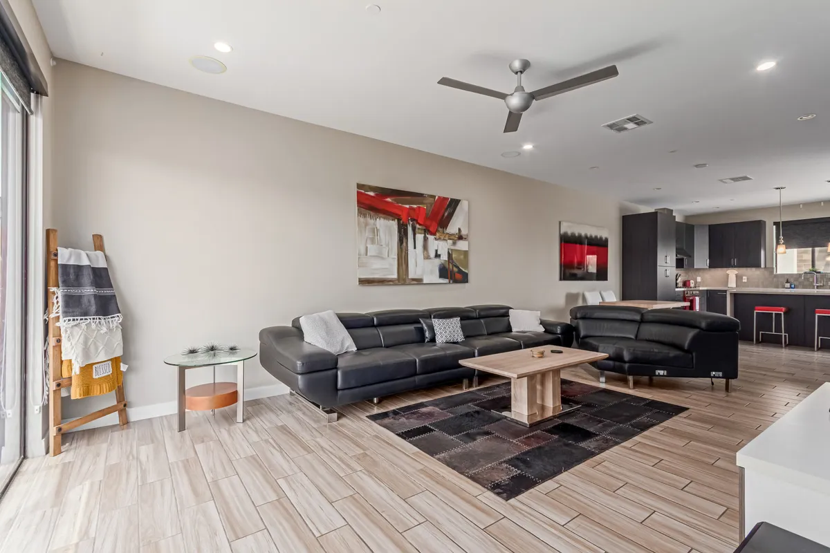 This inviting interior shot showcases a sunlit, open-concept living area with a sleek black leather sofa, a modern wooden coffee table, vibrant abstract art, and a stylish kitchen visible in the background. The room exudes contemporary comfort, with ample space for relaxing, entertaining, or unwinding after a day exploring Scottsdale.