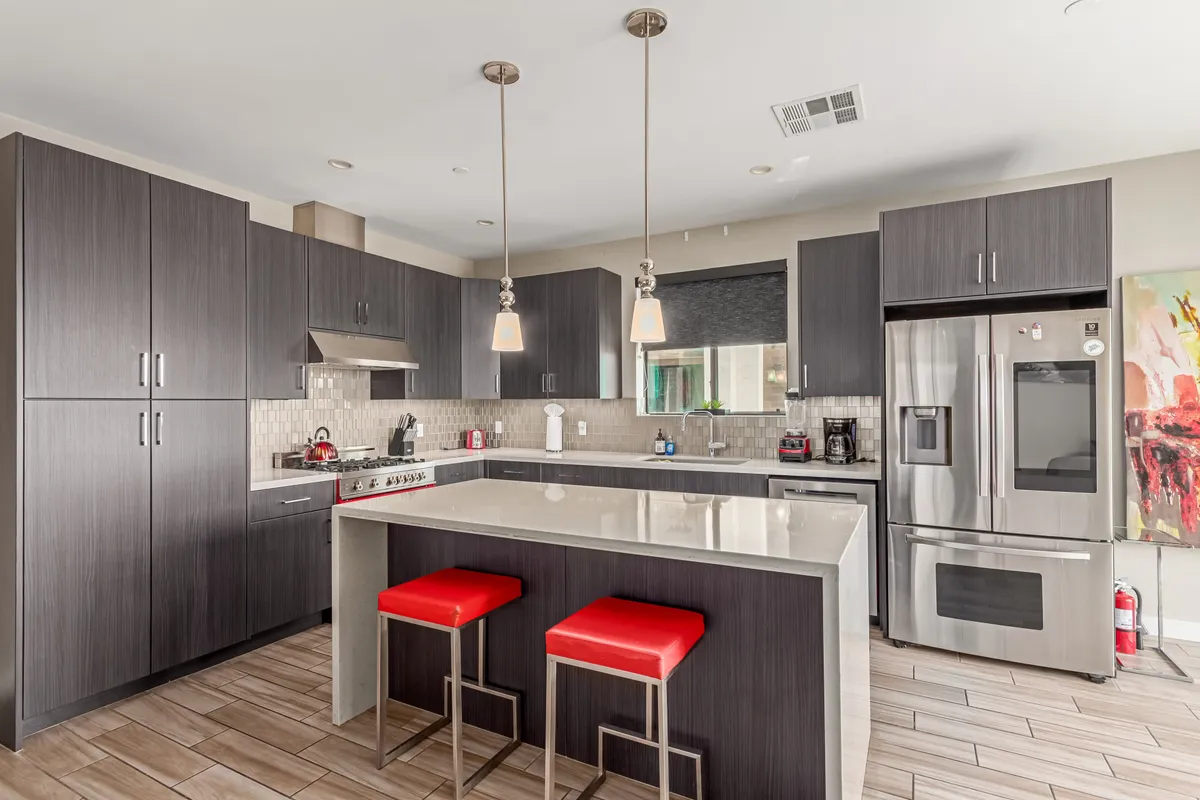 A stylish, contemporary kitchen featuring a prominent kitchen island with red bar stools, stainless steel appliances, and vibrant artwork on the wall. Ample natural light brightens the space, inviting both culinary creativity and social gatherings.