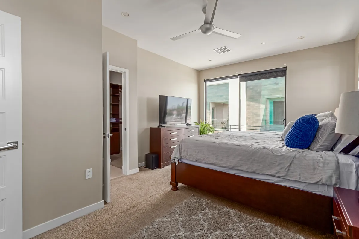 A bedroom with gray bedding, large TV on a dresser, decorative plant, and spacious windows that let in plenty of natural light. The decor is peaceful and minimal, creating a cozy retreat for relaxation or unwinding with a movie.