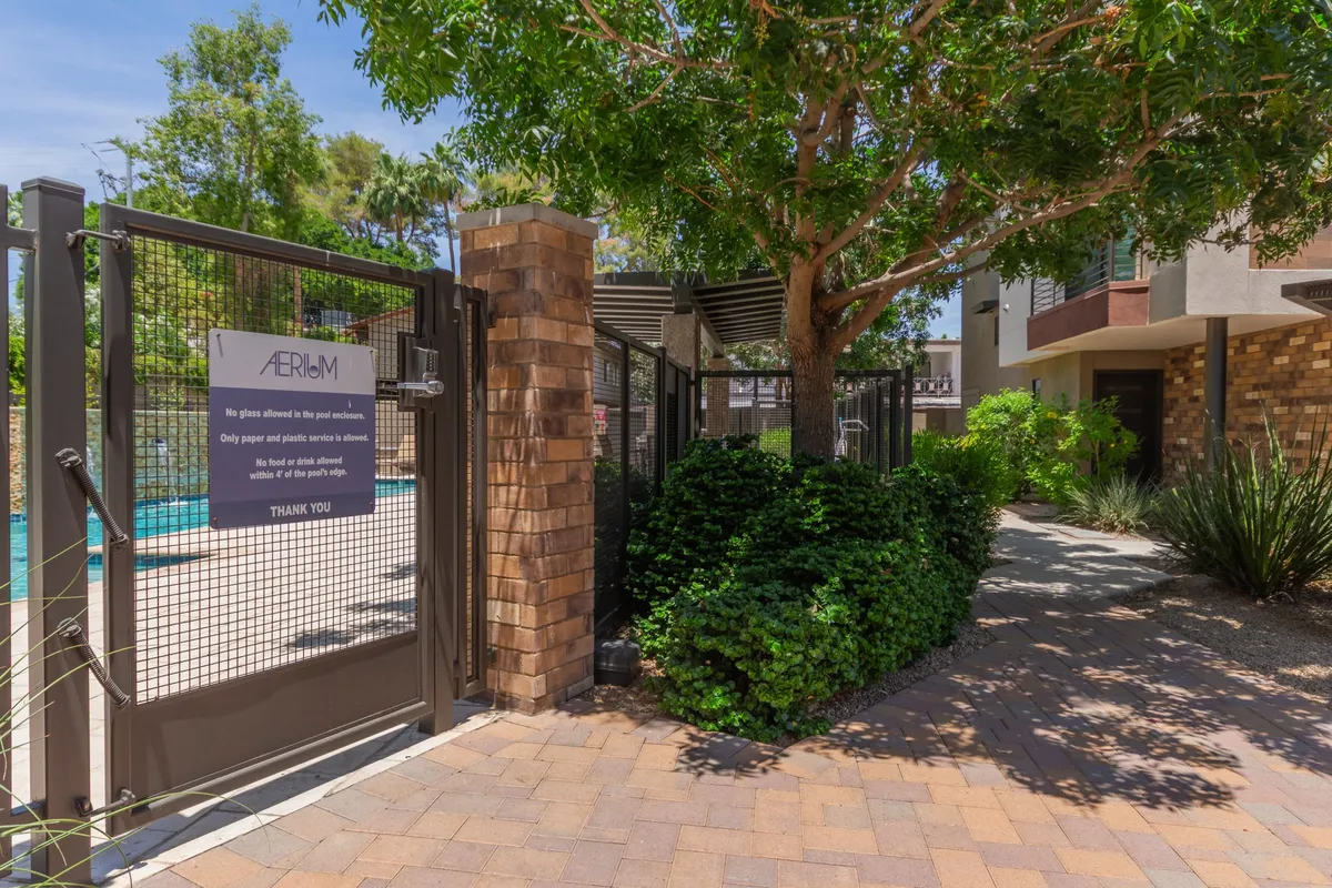 This exterior image features a welcoming gate entrance framed by lush greenery and brick pillars, with a glimpse of a serene pool area in the background. The inviting landscaping and open pathway create a sense of connection to the wider community and outdoor activities.