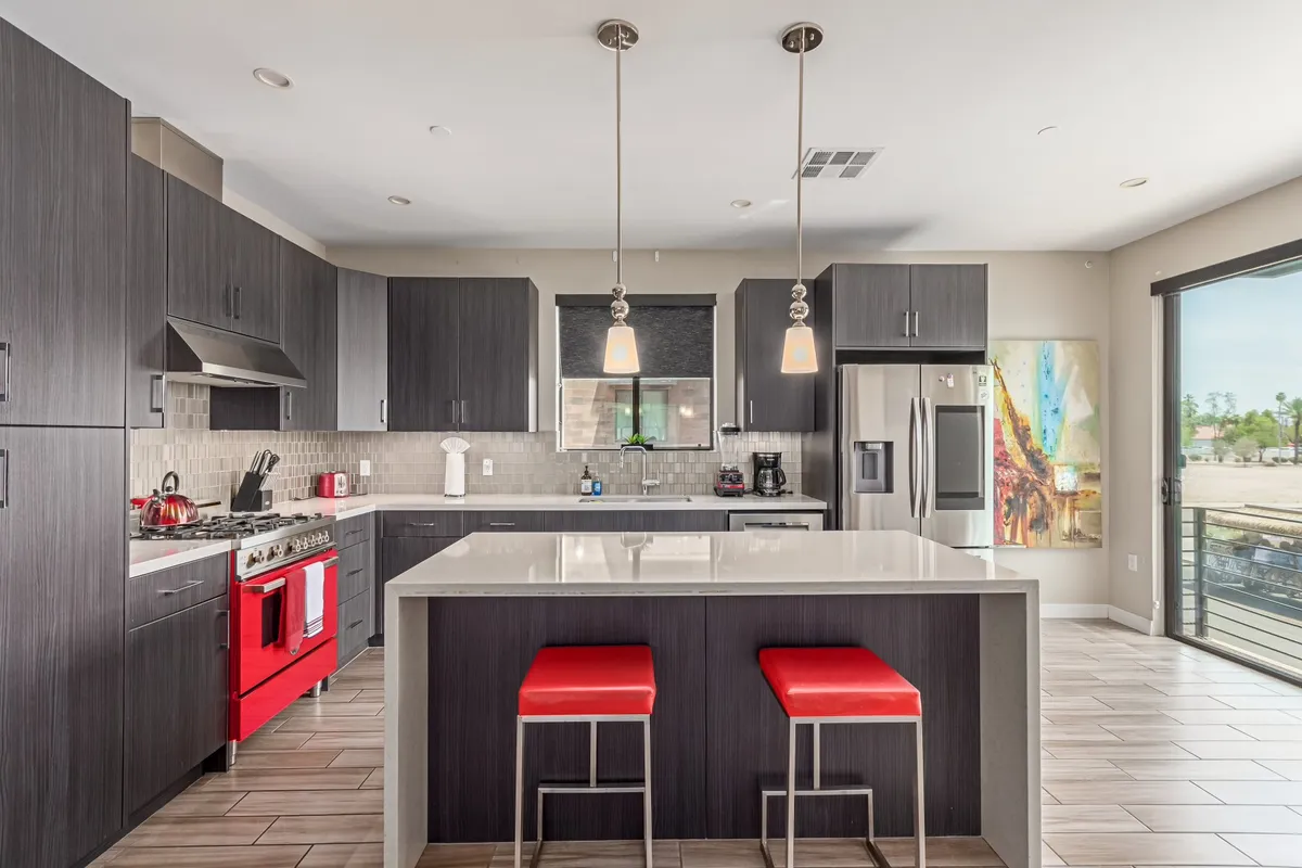 A vibrant kitchen setting featuring a bold red stove, white countertops, and matching red bar stools. Stylish artwork and abundant morning light complete the scene, making it perfect for both casual coffee and lively gatherings.