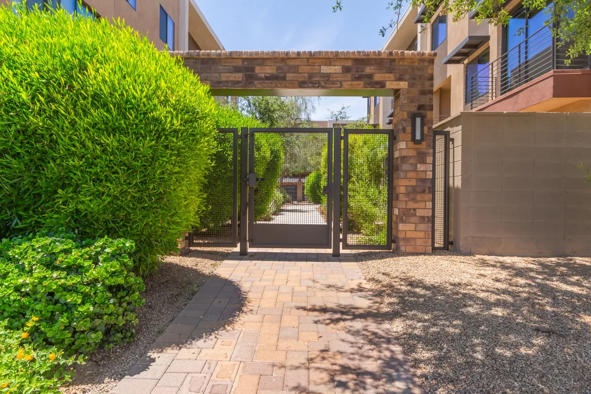 A welcoming pathway bordered by manicured hedges leads through a secure gate, offering a tranquil, garden-like setting ideal for leisurely walks or moments of relaxation. The scene captures the transition from the quiet, private community into the neighborhood, emphasizing a sense of safety and connection to the outdoors.